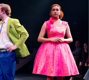 Onstage, a man in a lime green jacket points out of frame. Next to him, a woman stands in a hot pink dress with a cupcake-like skirt.