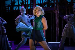 Onstage, a blonde woman whisks off her skirt to reveal blue shorts while people made to look like grey stone statues watch from behind.